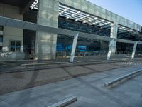 a bench sitting in front of a glass building on top of stone pavements below a metal ceiling