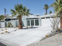 the house is surrounded by palm trees and desert rocks and gravel with a paved driveway leading to it