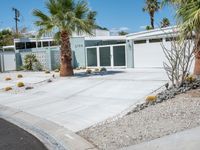 the house is surrounded by palm trees and desert rocks and gravel with a paved driveway leading to it