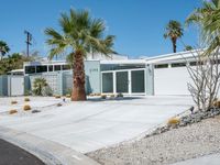 the house is surrounded by palm trees and desert rocks and gravel with a paved driveway leading to it