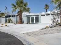 the house is surrounded by palm trees and desert rocks and gravel with a paved driveway leading to it