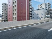 the side of an empty road next to a tall red building and a city with tall buildings