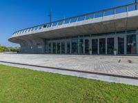 a concrete building with a lot of glass doors and windows, a red brick walkway and small grass and small green bushes in front