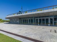 a concrete building with a lot of glass doors and windows, a red brick walkway and small grass and small green bushes in front