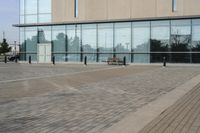 a glass building has benches on the sidewalk in front of it and cars parked in the driveway nearby