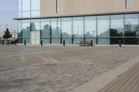 a glass building has benches on the sidewalk in front of it and cars parked in the driveway nearby