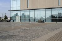 a glass building has benches on the sidewalk in front of it and cars parked in the driveway nearby