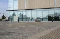 a glass building has benches on the sidewalk in front of it and cars parked in the driveway nearby