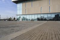 a glass building has benches on the sidewalk in front of it and cars parked in the driveway nearby