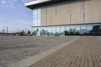 a glass building has benches on the sidewalk in front of it and cars parked in the driveway nearby