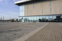 a glass building has benches on the sidewalk in front of it and cars parked in the driveway nearby