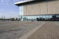 a glass building has benches on the sidewalk in front of it and cars parked in the driveway nearby