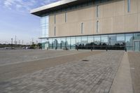 a glass building has benches on the sidewalk in front of it and cars parked in the driveway nearby