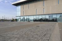 a glass building has benches on the sidewalk in front of it and cars parked in the driveway nearby