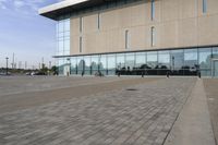 a glass building has benches on the sidewalk in front of it and cars parked in the driveway nearby