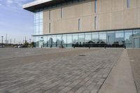 a glass building has benches on the sidewalk in front of it and cars parked in the driveway nearby