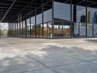 an empty walkway outside a glass building in the city with tables on the pavement and other architecture in the distance