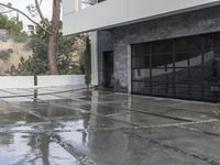 a glass garage is reflecting its surroundings on the wet ground outside with a white door and gray wall and windows