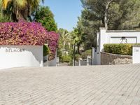 a driveway with trees, shrubs and a sign saying belmonto on it, with a small house with two cars behind it