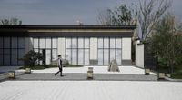 a woman walks in front of a modern house that is surrounded by white stone and plants