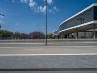 an empty street has a very big, glass - walled building in the background and trees along side