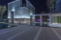 a modern building with an outdoor pool and a fenced area at night, on a sidewalk