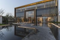 a long pool surrounded by a large glass building on top of a green hill behind it is two chairs