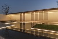 a reflecting pool in the middle of a building with lighted windows next to it and grass