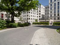 a cobblestone path between two residential buildings with trees and hedges in the foreground