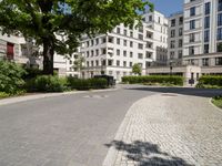 a cobblestone path between two residential buildings with trees and hedges in the foreground