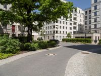 a cobblestone path between two residential buildings with trees and hedges in the foreground