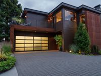 the driveway of this large home has black brick pavers in it, and lights are on