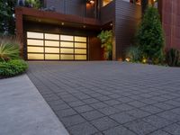 the driveway of this large home has black brick pavers in it, and lights are on