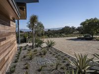 a home with plants on a gravel ground next to a building and mountains in the background