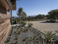 a home with plants on a gravel ground next to a building and mountains in the background