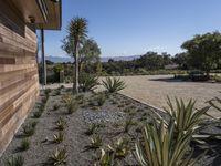 a home with plants on a gravel ground next to a building and mountains in the background