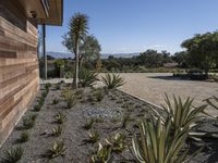 a home with plants on a gravel ground next to a building and mountains in the background