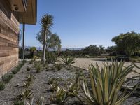 a home with plants on a gravel ground next to a building and mountains in the background