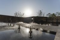 the reflection of the sun on the glassy surface of an outdoor pond in front of a wall with benches and umbrellas