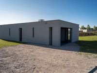 an empty gray house sitting on top of a gravel lot near a grassy area and a building