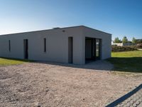 an empty gray house sitting on top of a gravel lot near a grassy area and a building