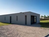 an empty gray house sitting on top of a gravel lot near a grassy area and a building