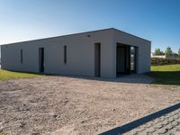 an empty gray house sitting on top of a gravel lot near a grassy area and a building