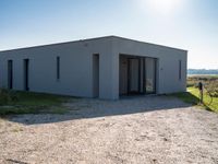 an empty gray house sitting on top of a gravel lot near a grassy area and a building