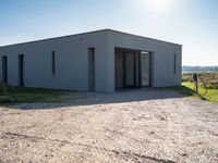 an empty gray house sitting on top of a gravel lot near a grassy area and a building