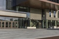 a modern style building with glass walls and entrance and sidewalk, and a few benches sitting on the front side, and in front of it