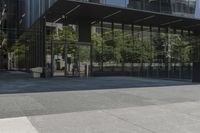 an empty bench is outside a glass building with trees reflected on the side of it