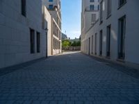 two people walking down an empty city street in an alley area with tall buildings in the background