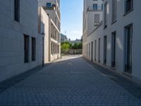 two people walking down an empty city street in an alley area with tall buildings in the background