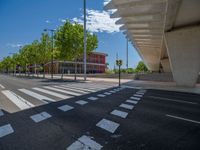 a car sitting at an empty street underneath a overpass overhang that contains a train stop, and cars and a pedestrian